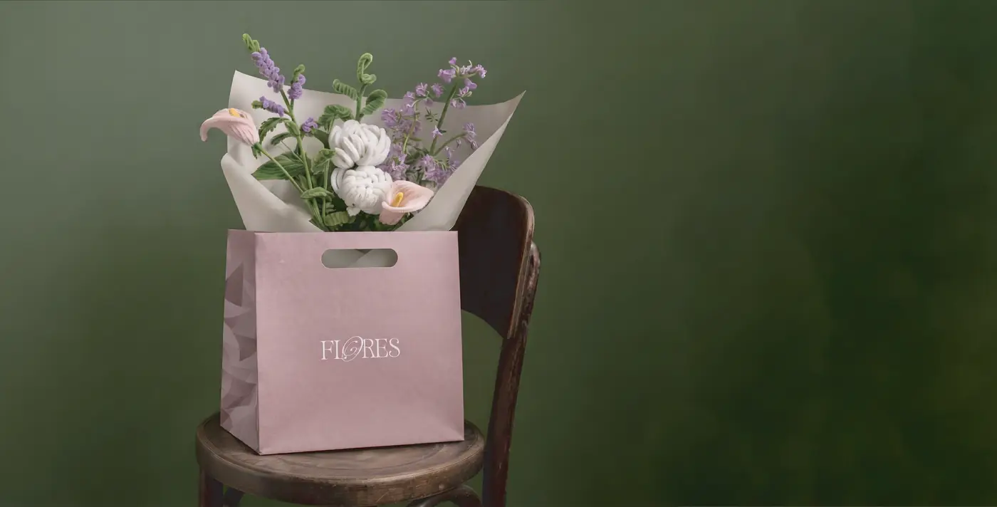 A Felores bouquet of calla lilies, lavender and white chrysanthemums in a blush gift bag, resting on a wooden chair